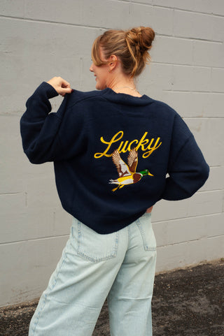 A woman wearing a navy blue sweatshirt with a yellow 'Lucky' script and a mallard duck graphic on the back, paired with light-wash jeans, standing against a gray brick wall.