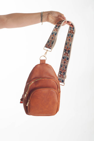 A small brown leather crossbody bag with a patterned fabric shoulder strap, held by a person's hand against a plain white background.
