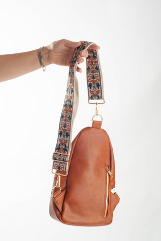 A tan leather crossbody bag with a wide, patterned fabric shoulder strap held up by a person's hand against a plain white background.