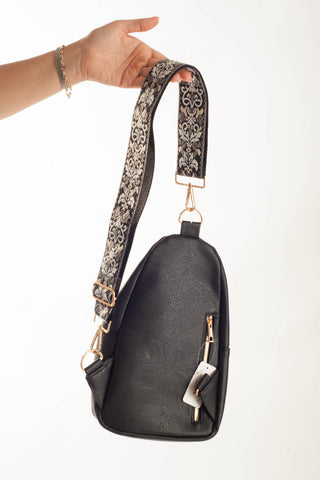 A black faux-leather shoulder bag with a wide, patterned black-and-beige strap and gold-tone hardware, held by a hand against a white background.