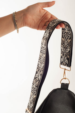 A hand holding a black handbag with a wide, patterned black and beige damask shoulder strap, featuring gold-tone hardware against a white background.