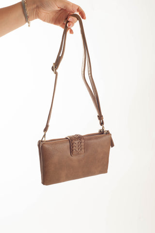 A brown faux-leather crossbody clutch bag with a woven detail on the front flap, held by a hand against a plain white background.