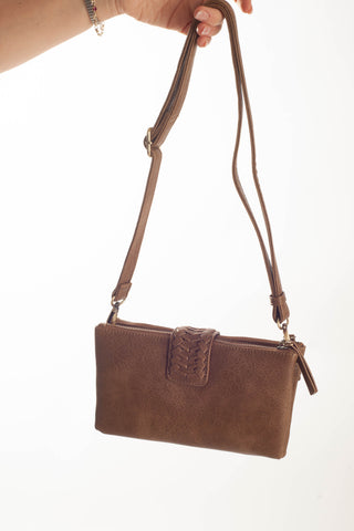 A brown faux-leather crossbody handbag with a braided front detail and adjustable shoulder strap, held by a hand against a plain white background.