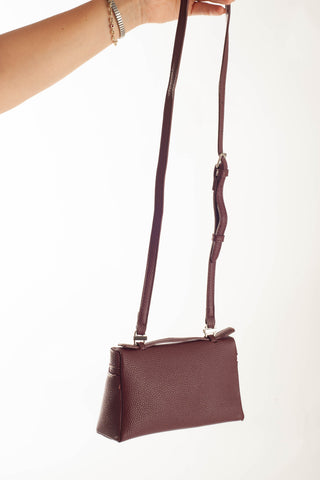 A burgundy pebbled leather crossbody bag with a long adjustable strap, held by a person's arm against a plain white background.