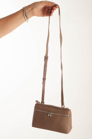 A taupe brown pebbled leather shoulder bag with a long adjustable strap, held by a person's hand against a plain white background.