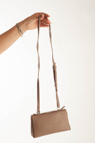A taupe pebbled leather crossbody bag with a matching adjustable shoulder strap, held by a person's hand against a plain white background.