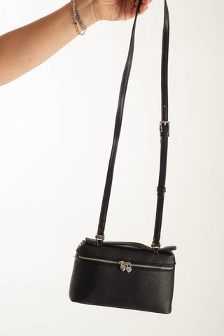 A black pebbled leather crossbody bag with a silver-tone double-bow clasp and a long adjustable shoulder strap, held by a person's hand against a plain white background.