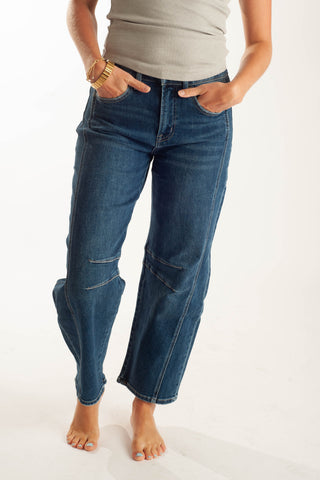 A person wearing dark blue cropped jeans with contrast light stitching and patch pockets, paired with a light gray sleeveless top, standing barefoot against a white studio background.