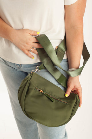 An olive green crescent-shaped crossbody bag with a wide matching shoulder strap, shown being worn by a person in a white t-shirt and light blue jeans.