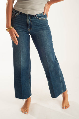 A person wearing dark blue cropped flare jeans with a raw hem, paired with a light gray top, shown against a plain white studio background.