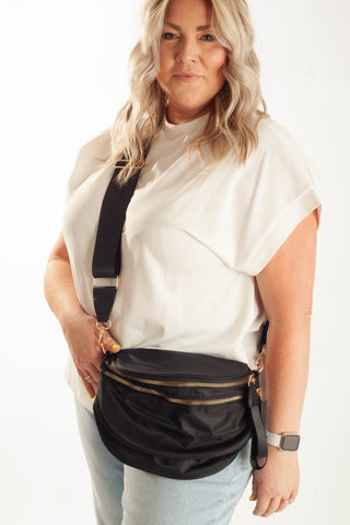 A woman wearing a white t-shirt and light blue jeans models a black nylon crossbody fanny pack with gold-tone hardware against a plain white background.