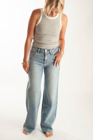 A woman wearing light-wash, wide-leg blue jeans and a gray ribbed tank top, standing against a plain white studio background.