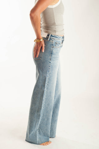 A person shown from the waist down wearing light-wash blue wide-leg jeans, a light gray tank top, and a gold bracelet, standing against a plain white studio background.