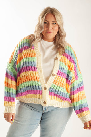 A woman wearing a chunky knit cardigan with horizontal rainbow stripes in yellow, orange, green, blue, pink, and purple over a white t-shirt and light blue jeans, photographed against a plain white studio background.