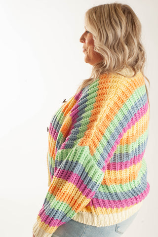 A woman shown from the side wearing a chunky knit cardigan with horizontal rainbow stripes in yellow, orange, pink, purple, blue, and green, paired with light blue jeans against a plain white background.