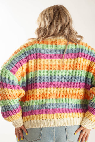 A woman viewed from behind wearing a chunky knit sweater with horizontal rainbow stripes in yellow, orange, pink, green, blue, and cream, paired with light-wash jeans against a plain white background.