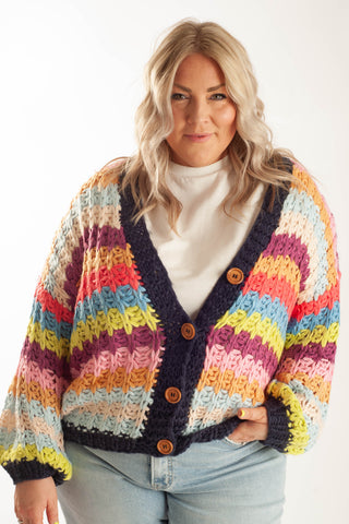 A woman wearing a chunky, multicolored striped crochet cardigan with a dark navy blue trim and brown buttons, paired with a white turtleneck and light blue jeans, against a plain white background.