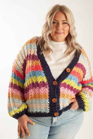 A woman wearing a chunky, multicolored striped crochet cardigan with brown buttons over a white turtleneck and light blue jeans, photographed against a clean white studio background.