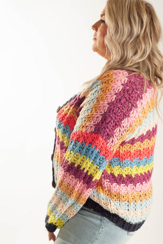 A woman shown from the side wearing a colorful, chunky knit cardigan with horizontal stripes in pink, orange, yellow, blue, and white, paired with light-wash jeans against a plain white background.