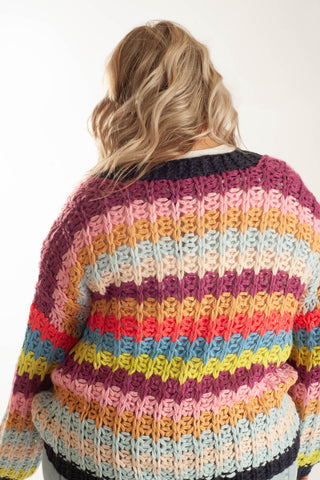 A woman viewed from behind wearing a chunky, hand-knit sweater with horizontal stripes in pink, orange, yellow, light blue, and purple, set against a plain white background.