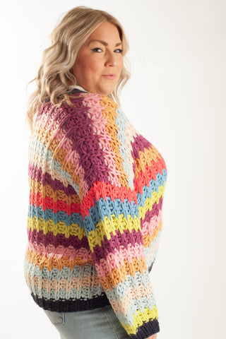 A woman wearing a chunky, hand-crocheted knit sweater with horizontal rainbow stripes in pink, purple, orange, yellow, blue, and green, shown from a three-quarter back view against a plain white studio background.