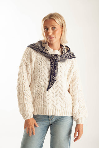 A woman wearing a cream cable-knit sweater with a navy and white patterned neck scarf, paired with light blue jeans, against a plain white studio background.