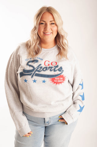 A woman wearing a light heather gray crewneck sweatshirt with a patriotic sports graphic that reads 'Let's Loud Go Sports' and 'Here for the snacks' with blue stars and blue sleeve graphics, paired with light blue jeans against a plain white background.