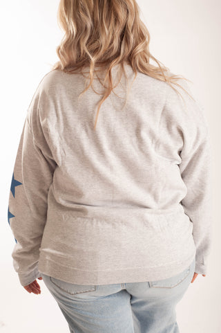 A woman viewed from behind wearing a light heather gray long-sleeve sweatshirt with blue star graphics on the sleeve, paired with light blue jeans against a plain white background.