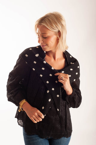 A woman with blonde hair wearing a black textured cardigan with white embroidered dots, paired with blue jeans, photographed against a plain white studio background.