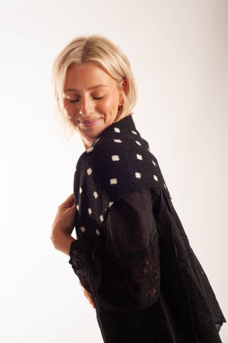 A blonde woman wearing a black knit wrap or shawl with white polka dots over a sheer black lace top, photographed against a plain white studio background.