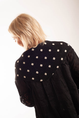 A woman with blonde hair is shown from behind wearing a black eyelet blouse with a contrasting black wool collar decorated with small white embroidered squares, set against a plain white background.