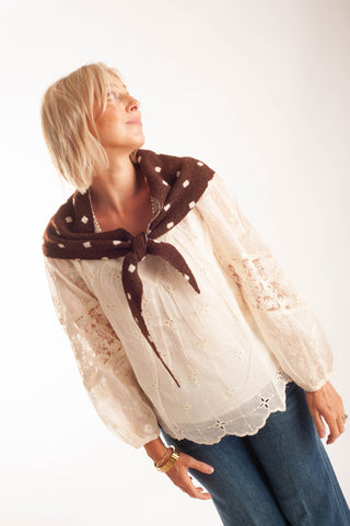 A woman wearing a cream lace blouse with a brown polka-dot scarf tied around her neck, paired with blue jeans, photographed against a plain white studio background.