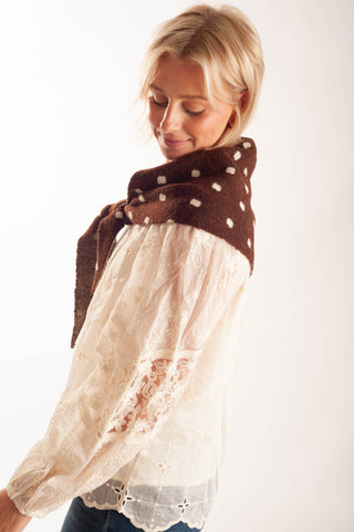 A woman wearing a brown knit shawl with white polka dots tied around her neck over a cream lace blouse, photographed against a plain white studio background.