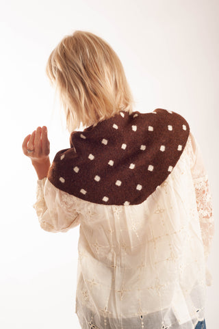 A woman viewed from behind wearing a brown woolen shawl with white polka dots over a cream lace blouse, set against a plain white background.