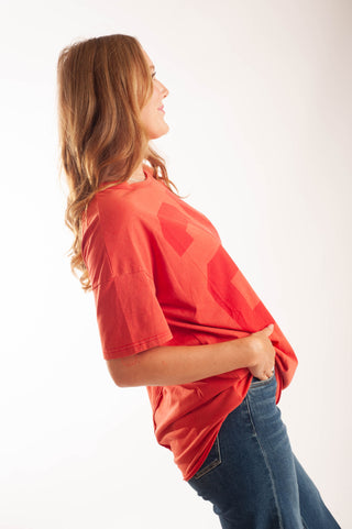 A woman in profile wearing a loose red-orange t-shirt with a tonal geometric pattern and blue jeans, photographed against a clean white studio background.