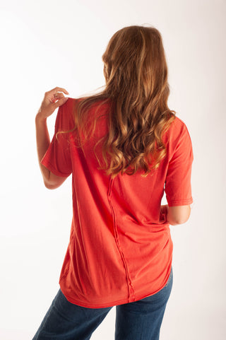 A woman with long wavy light brown hair is shown from behind wearing a bright red short-sleeve t-shirt and blue jeans against a plain white studio background.