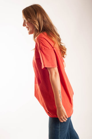 A woman with long light brown hair is shown in profile wearing an oversized short-sleeve t-shirt in a vibrant coral-red color, paired with blue jeans, against a plain white studio background.