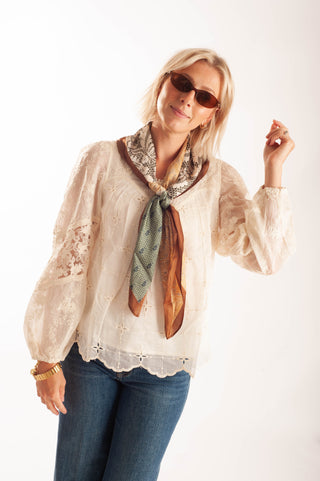 A woman modeling a cream lace blouse with a brown and green patterned scarf tied around the collar, paired with blue jeans and sunglasses, against a plain white background.