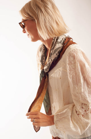 A woman with short blonde hair and sunglasses, wearing a cream lace blouse and a patterned scarf with brown and green tones, shown in profile against a plain white background.
