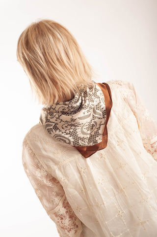 A woman with blonde hair is shown from behind, wearing a cream lace blouse and a patterned scarf with brown trim around her neck, set against a plain white background.