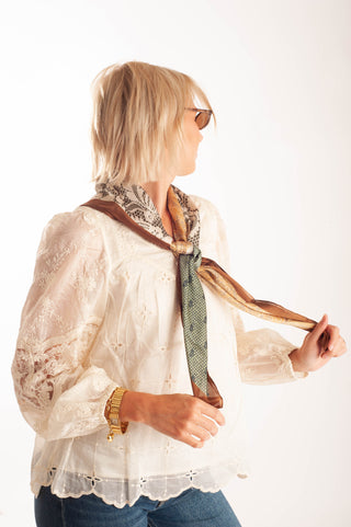 A woman with blonde hair and sunglasses wearing a cream lace blouse and blue jeans, modeling a brown and green patterned neck scarf against a plain white background.