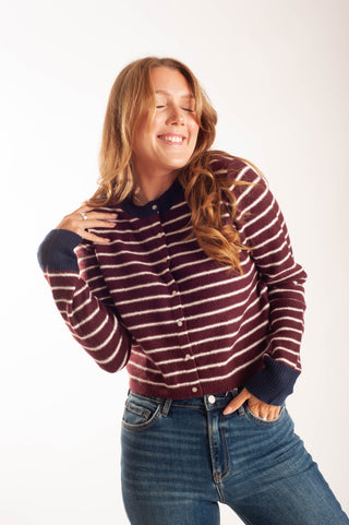 A woman with long light brown hair is smiling and posing in a burgundy and white striped knit cardigan with navy blue trim, paired with blue jeans, against a plain white studio background.