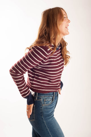 A woman with long light brown hair is shown in profile, wearing a maroon and white horizontally striped knit sweater with dark navy cuffs and collar, paired with blue jeans, against a plain white studio background.
