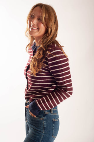 A woman with long wavy light brown hair is shown in profile, wearing a maroon and cream striped knit cardigan over a blue shirt and blue jeans, against a plain white studio background.