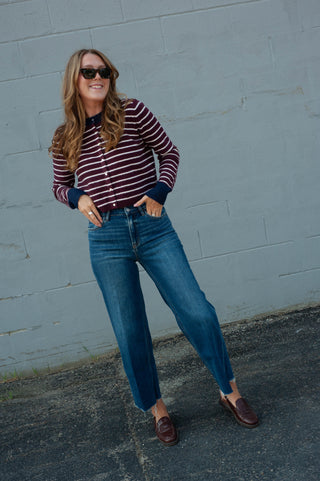 A woman wearing a burgundy and navy striped long-sleeve button-up shirt, blue jeans, and brown leather loafers, standing against a gray painted brick wall.