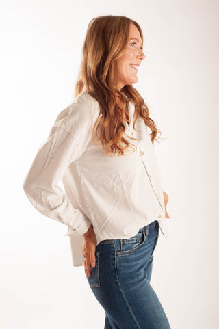 A woman with long wavy light brown hair is shown in profile wearing a cream-colored, long-sleeve button-up shirt and blue jeans against a plain white studio background.