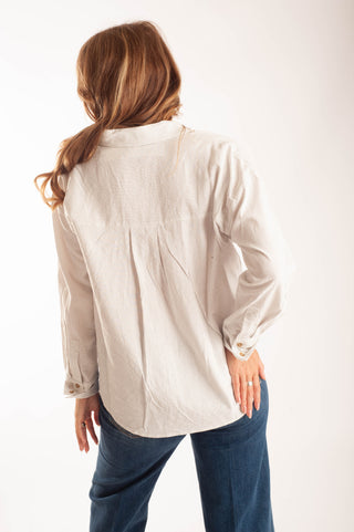 A woman viewed from behind wearing a light beige linen button-up shirt and blue jeans, standing against a plain white studio background.
