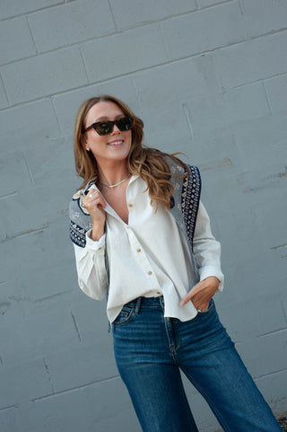 A woman wearing a white button-down shirt with dark patterned shoulder details, blue jeans, and sunglasses, posing against a gray painted brick wall.