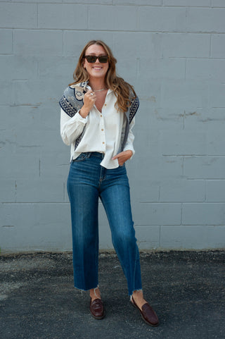 A woman stands outdoors against a gray painted brick wall wearing a white long-sleeve button-up shirt with dark embroidered shoulder details, blue wide-leg jeans, and dark brown loafers.