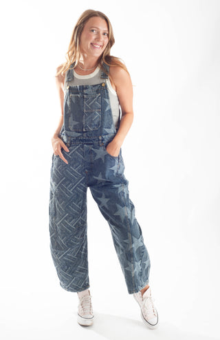 A woman modeling a pair of blue denim overall-style pants with a faded geometric and star pattern, worn over a white tank top and white sneakers against a plain white studio background.